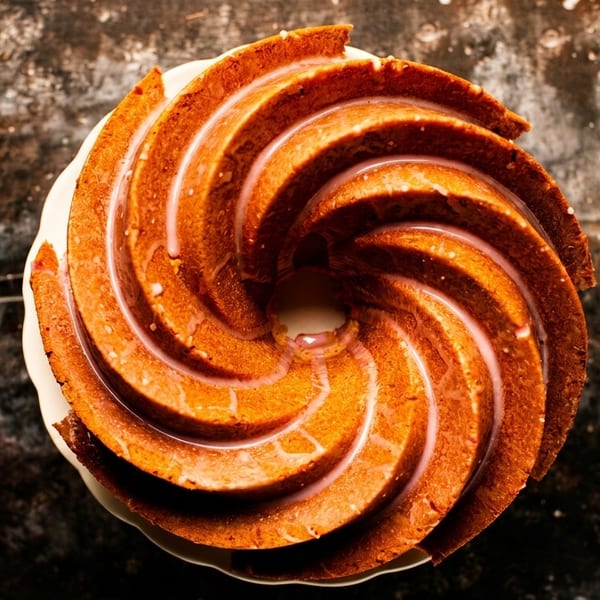 Overhead view of a golden-brown bundt cake with a light pink glaze, on a white plate.